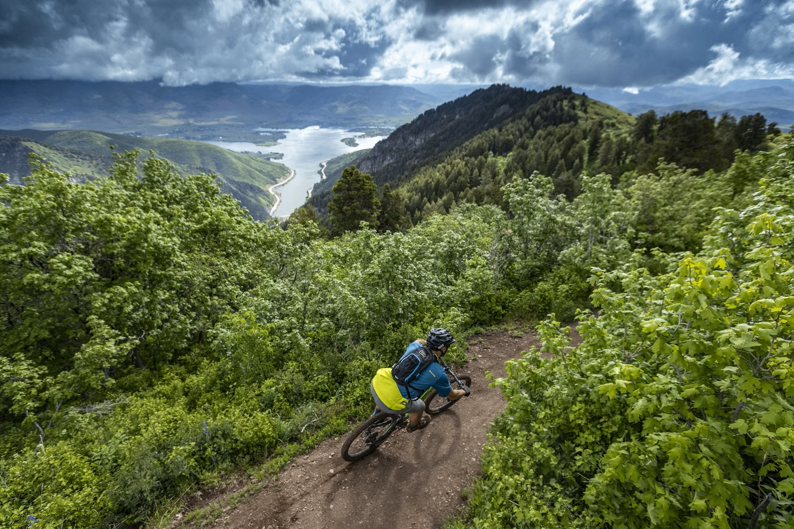 Mountain biker on a dirt trail above Pineview Reservoir with green hills and storm clouds beyond