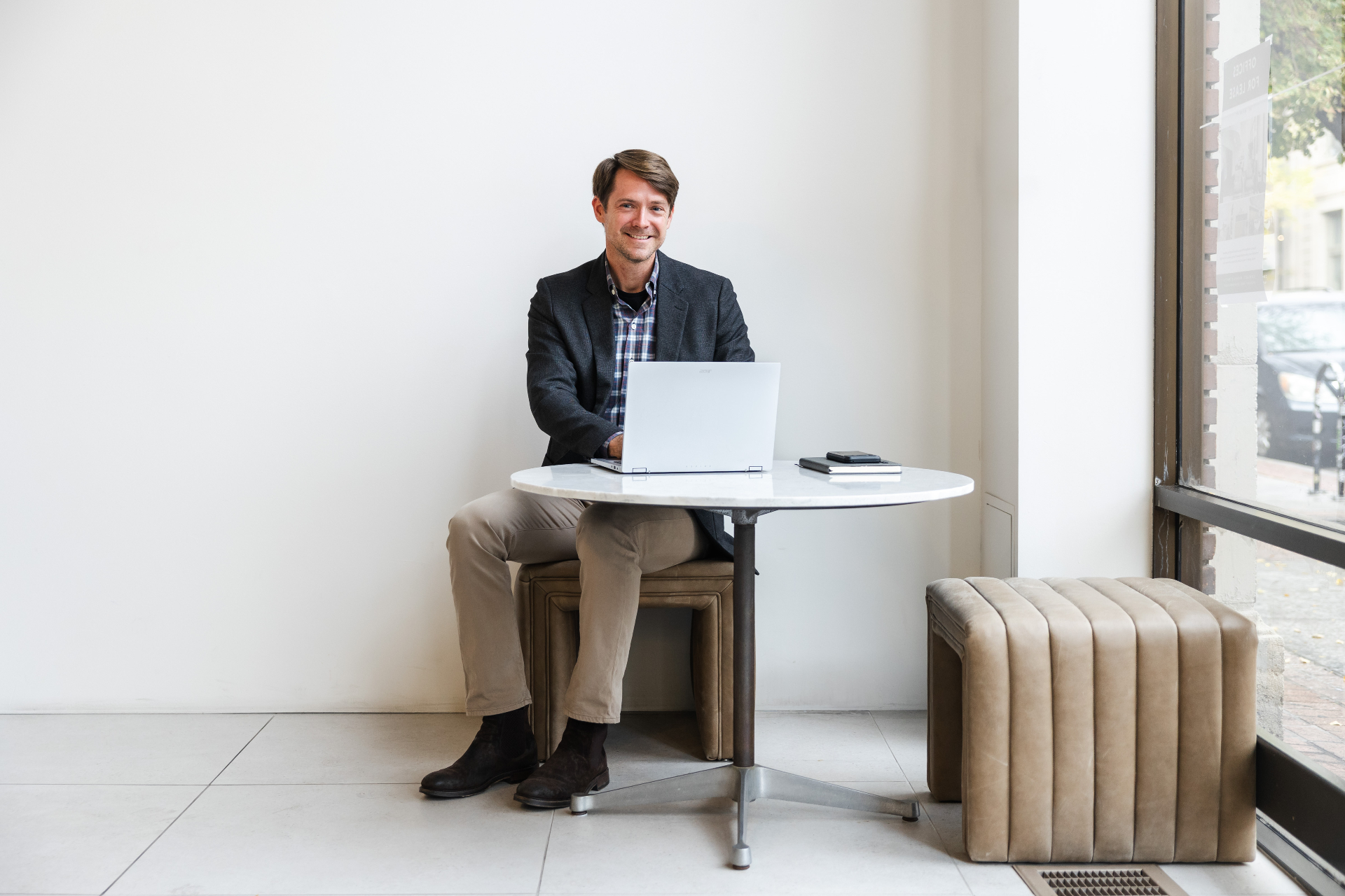 Patten Wood seated at a round marble table with a laptop, smiling toward the camera in a bright storefront space